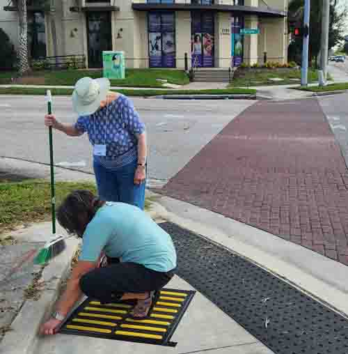 picture shows two women st sn intersection, one of them is sqatting with her hands on a black and white striped detectable tile.